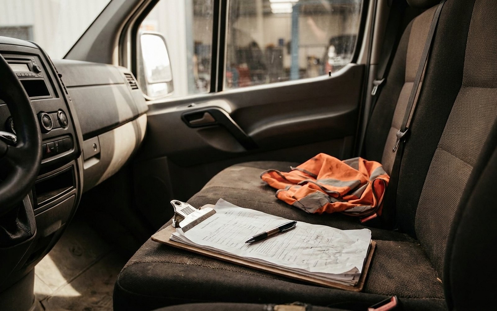 Klemmbrett mit Formular und Stift auf dem Beifahrersitz eines Transporters, daneben eine orange Warnweste